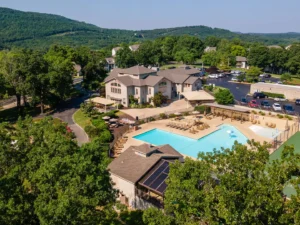 Aerial view of Pointe Royal Branson MO resort with a large outdoor swimming pool, lounge chairs, umbrellas, and surrounding trees, set against green hills and a clear sky—perfect for relaxing Branson vacation rentals.