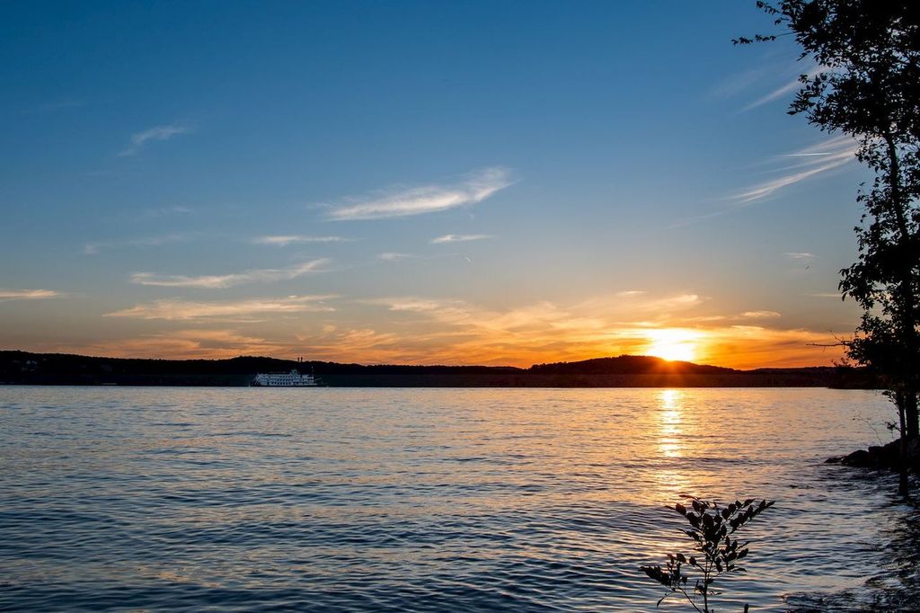 A serene sunset over a calm lake with gentle ripples, a cruise ship in the distance, silhouetted hills on the horizon, and trees framing the scene—an ideal setting for Branson vacation rentals.