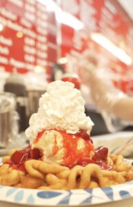 A plate of funnel cake topped with vanilla ice cream, strawberry sauce, and whipped cream. Blurred red menu in the background enhances the indulgent feel.