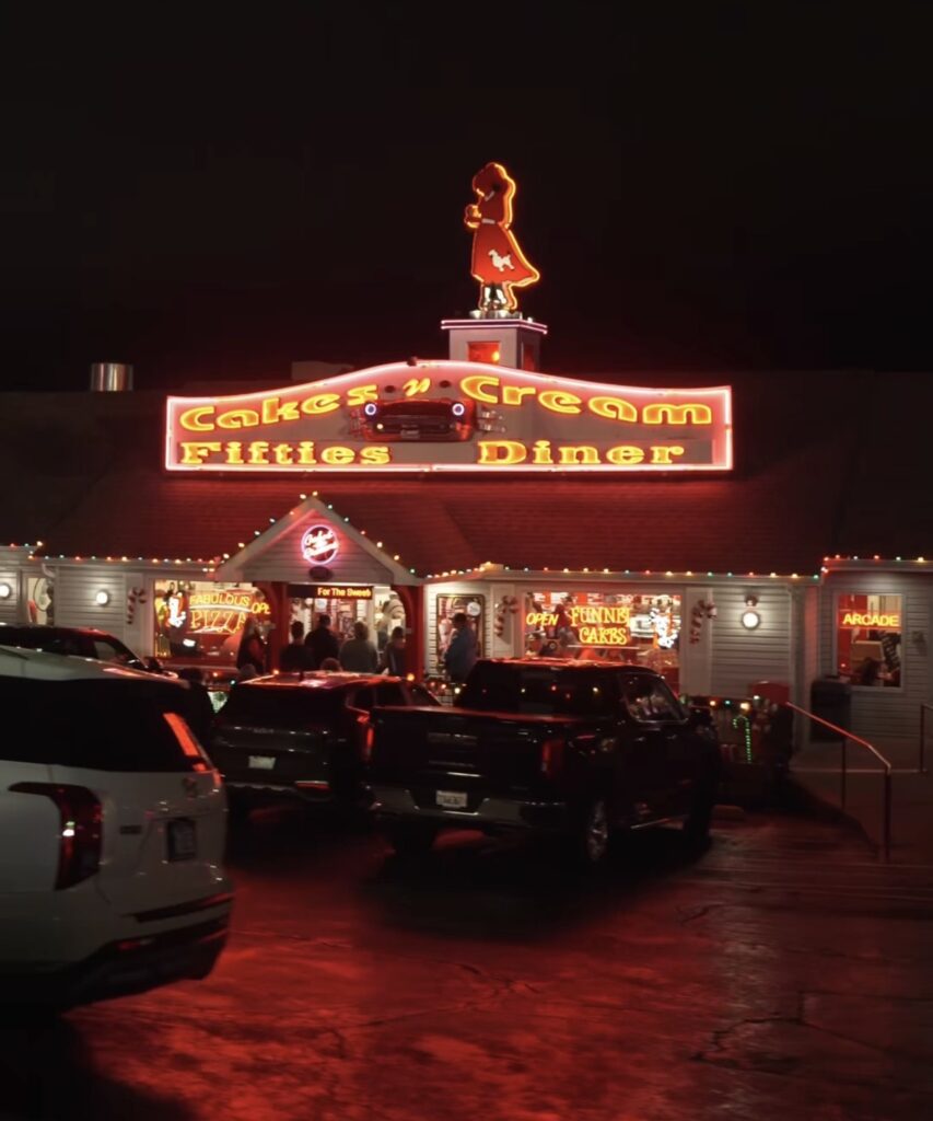 Exterior night view of a 1950s-style diner with neon lights and signage reading "Cakes 'n' Cream Fifties Diner." Several cars are parked outside, and people are visible through large windows. The atmosphere is nostalgic and lively.