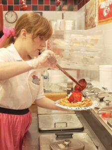 Staff member preparing a fresh funnel cake topped with ice cream and strawberry sauce at Cakes N Creams.
