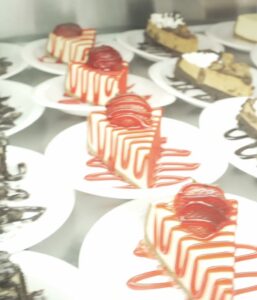 Slices of strawberry cheesecake drizzled with red sauce on white plates inside the dessert case at Cakes N Creams.