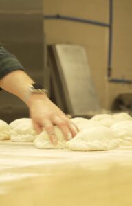 A simply baked employee gently arranges raw dough pieces on a floured table in a bakery setting. The scene conveys a sense of precision and craftsmanship.