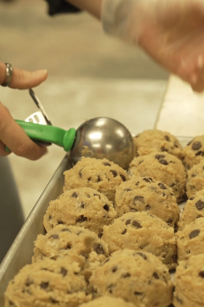 Close-up of hands using a green scoop to portion simply baked chocolate chip cookie dough onto a baking tray. The scene conveys anticipation and warmth.