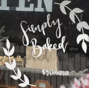 Storefront window displaying "Simply Baked by Chandler" in white script with leaf design and bold "OPEN" sign, evoking a welcoming bakery vibe.