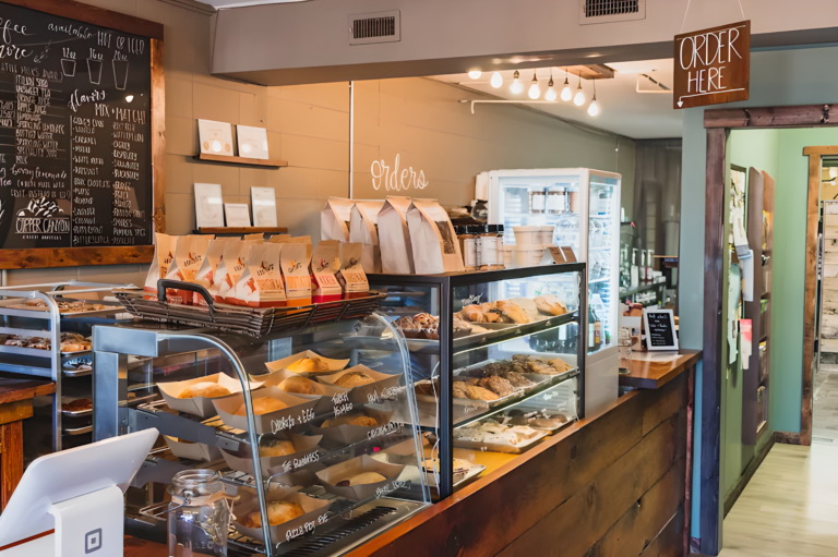 Simply Baked's Cozy café interior with a wooden counter displaying an array of pastries and snacks. A chalkboard menu and "Order Here" sign add a welcoming feel.