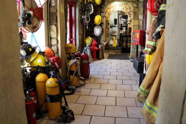 A hallway lined with fire extinguishers, oxygen tanks, fire helmets, and jackets leads to a room filled with firefighting memorabilia. Stone walls and tiled floors create a museum-like setting inside Hook and Ladder Pizza.