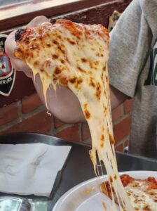 A person lifts a cheesy slice of Hook and Ladder Pizza from a plate, melted cheese stretching between the slice and the rest of the pizza. The background features a brick wall and part of the person’s gray shirt.