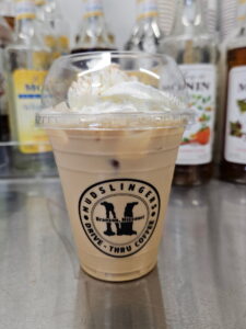 A plastic cup of iced coffee from Mudslingers, topped with whipped cream, sits on a counter. Various syrup bottles are blurred in the background.