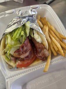 The Philly Truck gyro wrapped in foil with bacon, lettuce, tomato, and sauce in a pita, served with golden fries in a white foam takeout container.