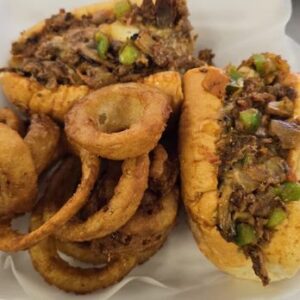 A Philly Truck cheesesteak loaded with beef, green peppers, and onions, served with crispy, golden brown onion rings on a white paper-lined basket.