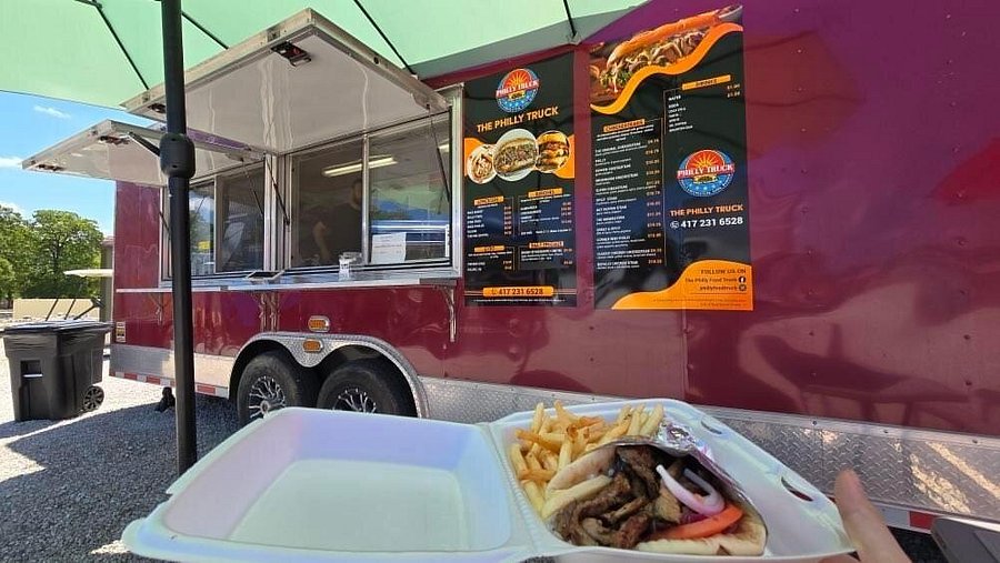 The Philly Truck located under a sunny sky, offering Philly cheesesteaks, with a menu displayed. In the foreground, a person holds a box with a sandwich and fries.