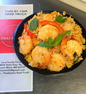 A Thai by Tom black bowl filled with shrimp fried rice, garnished with basil leaves and red bell peppers. A menu from "Thai by Tom Food Truck" is partially visible.