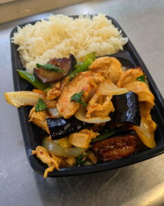 Thai by Tom rectangular black tray filled with stir-fried chicken, eggplant, onions, and basil. Jasmine rice is served beside it. The dish appears savory and appetizing.