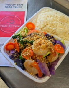 A vibrant Thai by Tom Thai dish featuring colorful vegetables, shrimp, and crispy garnish alongside steamed rice in a compartment tray. A Thai food truck menu is nearby.
