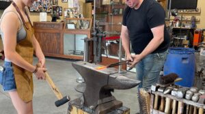 Silver Anvil Metalcraft employee in a black shirt and jeans uses tongs on hot metal at an anvil while another in a brown apron and denim shorts holds a hammer, inside a workshop.