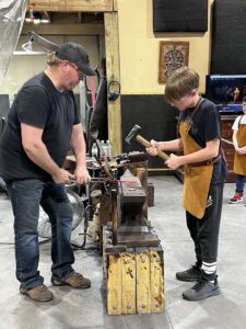 A man and a boy at Silver Anvil Metalcraft wearing aprons work at an anvil in a workshop, holding hammers poised to strike metal. The atmosphere is focused and instructional.