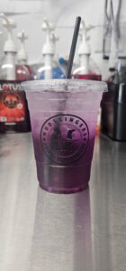 A plastic Mudslingers cup filled with a vibrant purple drink and a black straw, placed on a metallic counter. Bottles are blurred in the background, conveying a café setting.