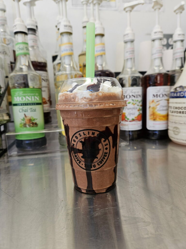 Mudslingers Iced chocolate drink in a clear cup with whipped cream and chocolate drizzle, placed on a metal counter. Syrup bottles line the background.