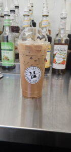 A tall Mudslingers iced coffee in a clear cup with a logo, filled with ice cubes, on a stainless steel counter. Several Monin syrup bottles are blurred in the background.