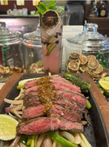A sizzling Little Hacienda plate of grilled steak with sauce, sliced lime, and vegetables. Behind it, a tall pink drink with a dried lime and mint garnish, conveying a fresh, appetizing scene.