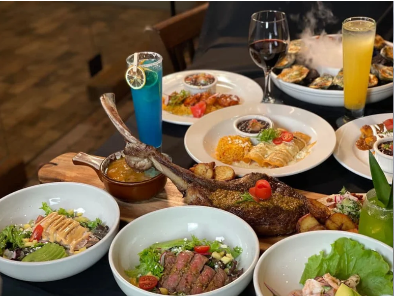 A lavish spread of Little Hacienda gourmet dishes, including a tomahawk steak, colorful salads, pasta, and seafood, with vibrant drinks and an elegant ambiance.