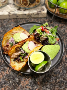 Two Little Hacienda tacos filled with meat, avocado sauce, radish, and onions on a black plate with a fresh green salad and lime wedges, set on a dark stone surface.