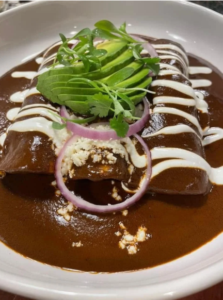 A plate of Little Hacienda enchiladas topped with rich mole sauce, drizzled with crema, garnished with sliced avocado, red onion rings, and cilantro, evoking a savory, festive feel.