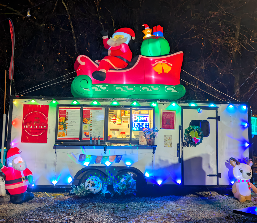 Thai by Tom food truck decorated with colorful lights and a large Santa inflatable on top. A lit "Open" sign is visible, creating a joyful, holiday ambiance.
