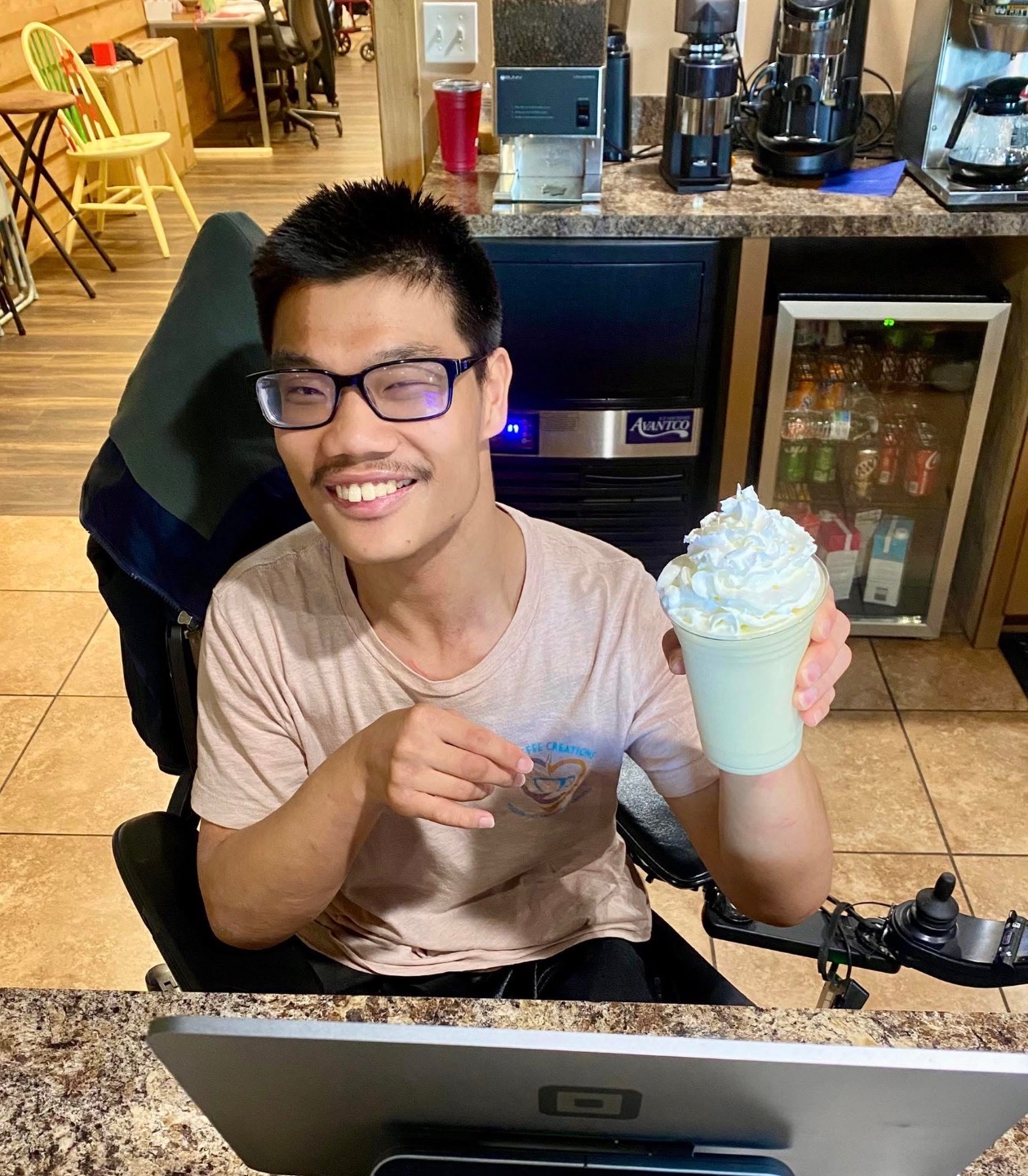 Coffee Creations employee smiling in a wheelchair holds a whipped cream-topped drink in a cozy cafe, radiating joy. Coffee machines and colorful chairs in the background.