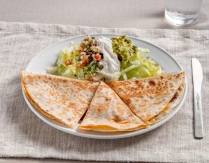 A plate of crispy quesadillas is served with shredded lettuce, guacamole, sour cream, and salsa on a beige cloth. A knife is placed beside the plate.