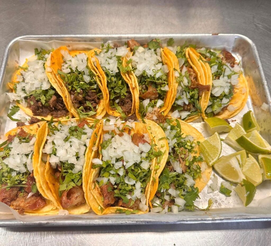 A tray of six tacos filled with chopped onions, cilantro, and meat, garnished with lime wedges. The setting conveys a fresh, appetizing tone.