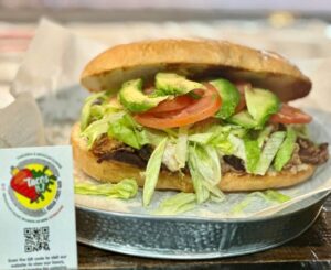 A fresh torta sandwich with layers of avocado, tomato, lettuce, and meat on a crusty bun sits on a tray. A restaurant card is displayed beside it.
