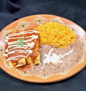 A vibrant plate features an enchilada topped with red sauce and white drizzle, served with orange rice and creamy refried beans, on a decorative ceramic dish.