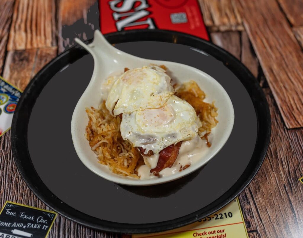 A hearty breakfast dish of hash browns topped with crispy bacon, creamy gravy, and two sunny-side-up eggs, served on a black plate. Rustic wood table underneath.