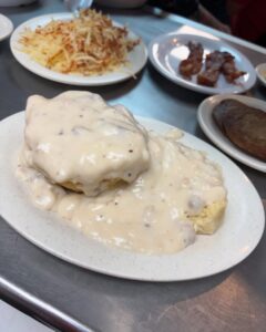 Biscuits topped with creamy white gravy on a plate, accompanied by hash browns, crispy bacon, and a sausage patty on a stainless steel table.