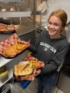 Smiling woman in a kitchen holds two baskets of food. One basket has a burger and curly fries, the other a sandwich and fries. Casual, cheerful atmosphere.