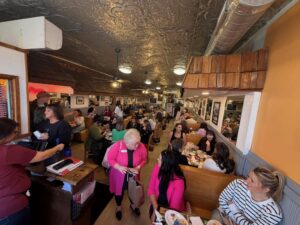A bustling restaurant scene with many people seated at wooden booths, engaged in conversation. Warm lighting creates a cozy, sociable atmosphere. A woman in a bright pink jacket stands, adding a pop of color. The ceiling has ornate tin detailing.