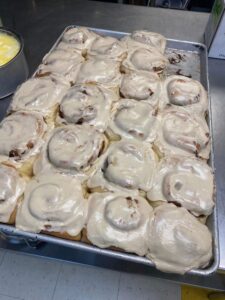 A tray of freshly baked cinnamon rolls topped with creamy icing, arranged in neat rows. The setting is a kitchen with a counter in the background.