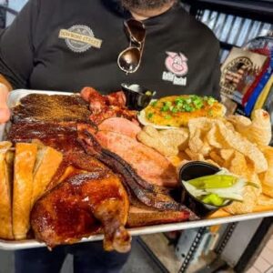 A person holds a tray of assorted barbecued meats, sliced bread, crispy pork rinds, and a side of veggies. The setting suggests a casual, hearty meal.