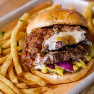 A juicy double cheeseburger with crispy onion rings, purple onions, and mustard, served with golden French fries on a tray, creating a savory and indulgent scene.