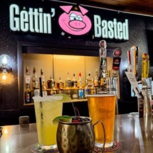 Bar counter at Gettin' Basted with three drinks: a green cocktail, a mug with lime, and a pint of beer; liquor bottles line the background.