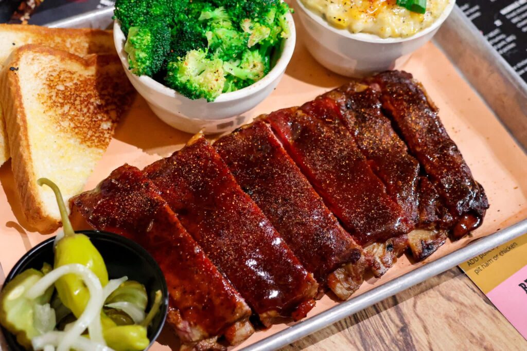 A tantalizing platter of glazed barbecue ribs with a side of vibrant steamed broccoli, creamy mac and cheese, toasted bread, and pickled peppers.