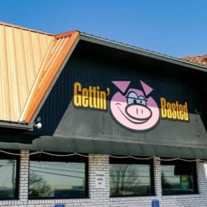 The image shows the exterior of a restaurant named "Gettin' Basted." The sign features a smiling pig logo on a black and white brick wall.