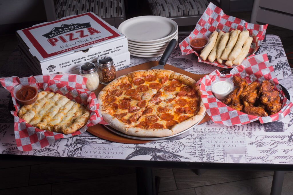 A pepperoni pizza on a wooden tray is surrounded by breadsticks, chicken wings, and cheesy bread, all on a red-checkered paper with dipping sauces.