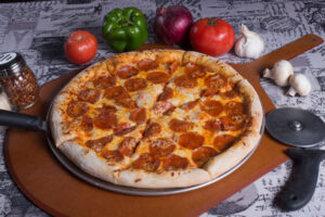 A pepperoni pizza on a wooden board, surrounded by fresh vegetables, garlic, mushrooms, and spices. A pizza cutter lies beside it, ready for serving.