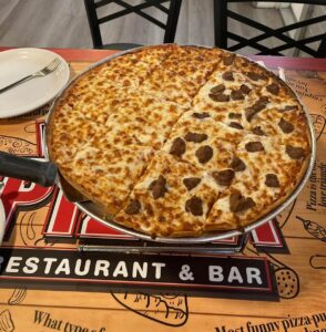 A thin-crust pizza with half cheese and half sausage topping, served on a metal tray atop a restaurant table with a white plate and fork nearby.