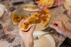 A close-up of a hand holding a slice of pizza with toppings like green peppers and sausage. Other slices are on a metal tray on a patterned tablecloth.