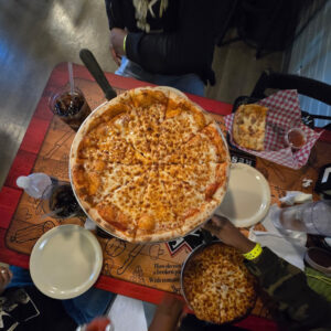 A table with a large cheese pizza at the center, surrounded by plates and drinks. A breadstick basket is nearby. The scene feels casual and inviting.