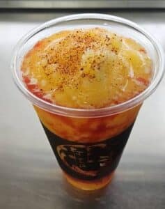 A tall plastic Bobalstic cup filled with vibrant mango smoothie topped with chili powder and syrup. The bright colors suggest a refreshing and spicy flavor blend.